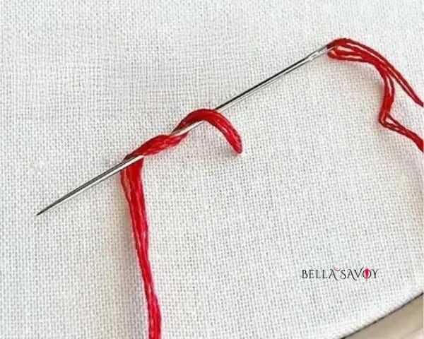 French Knot Tutorial 30 Close-up of how to do a French knot showing red embroidery floss wrapped twice around an embroidery needle on white fabric.