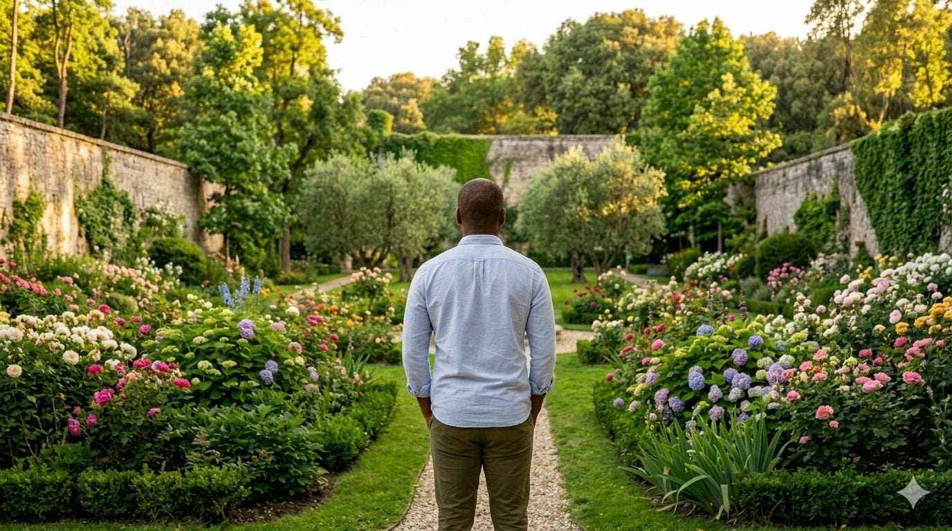 A Black man with closely cropped hair wearing a denim shirt and green pants stands in a colorful walled garden filled with flowers and trees
