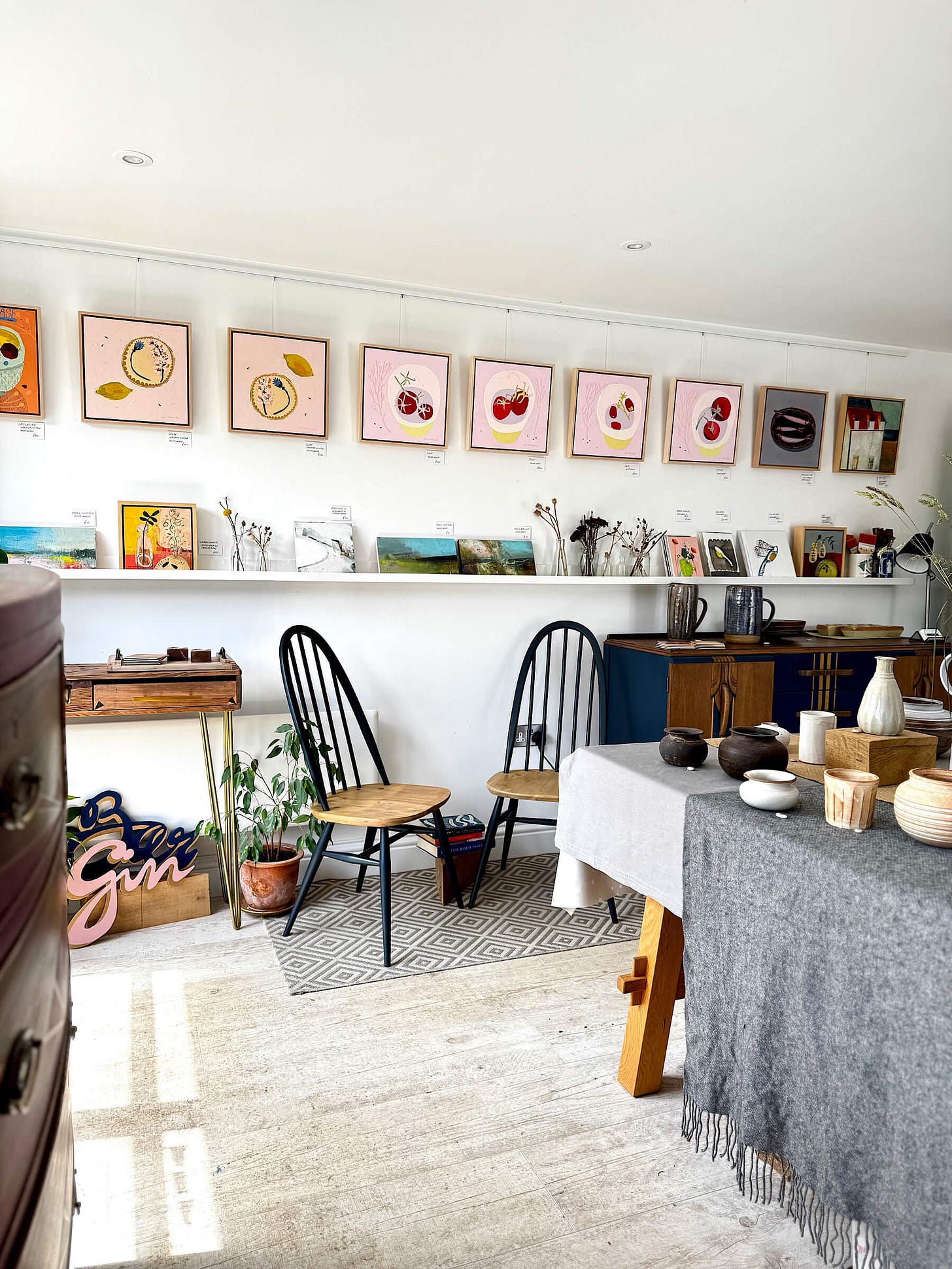 A bright, well-lit art studio interior showing a curated exhibition space called the Conversation Corner. A long white gallery wall features a row of framed mixed-media paintings on pink-primed backgrounds. Beneath the paintings, two elegant navy wooden spindle-back chairs sit on a geometric patterned rug next to a small wooden side table with gold legs. In the foreground, a large wooden table covered with grey and white tablecloths displays various handmade ceramic vessels, cups, and bowls. A bright, well-lit art studio interior showing a curated exhibition space called the Conversation Corner. A long white gallery wall features a row of framed mixed-media paintings on pink-primed backgrounds. Beneath the paintings, two elegant navy wooden spindle-back chairs sit on a geometric patterned rug next to a small wooden side table with gold legs. In the foreground, a large wooden table covered with grey and white tablecloths displays various handmade ceramic vessels, cups, and bowls.