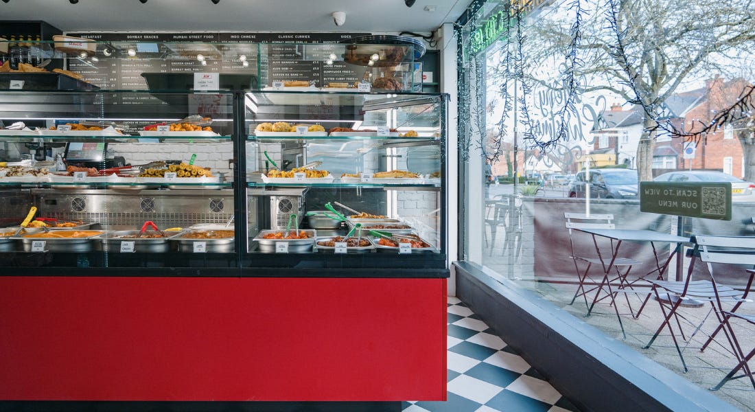 A shop with a red counter with Indian food inside of it, menus on the wall, and glass windows with tables outside A shop with a red counter with Indian food inside of it, menus on the wall, and glass windows with tables outside