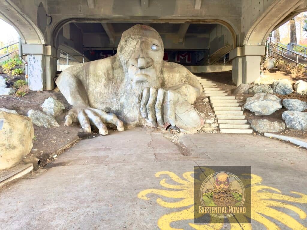  This is a photograph of a large, concrete sculpture of a troll, known as the Fremont Troll, located under a bridge. The troll appears to be hunched over, holding an actual Volkswagen Beetle. Its left eye is a hubcap. The ground in front of the troll is paved, and a yellow, sun-like design is painted on the pavement. There are stairs and large rocks on the right side of the image, and a brick wall with graffiti is visible in the background under the bridge's archway.