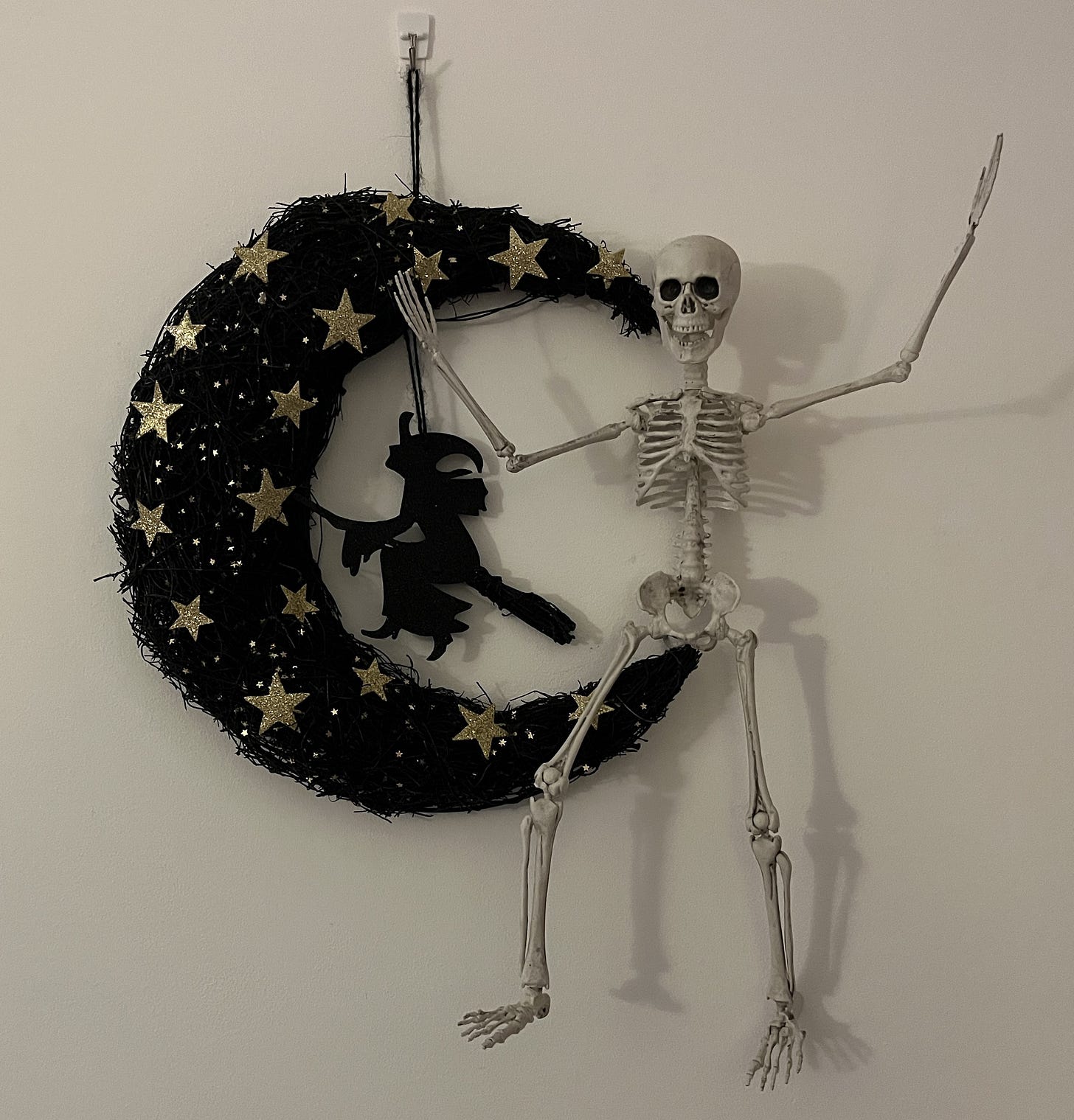 A toy skeleton sitting on a hanging decoration of a black crescent moon with a witch on it.