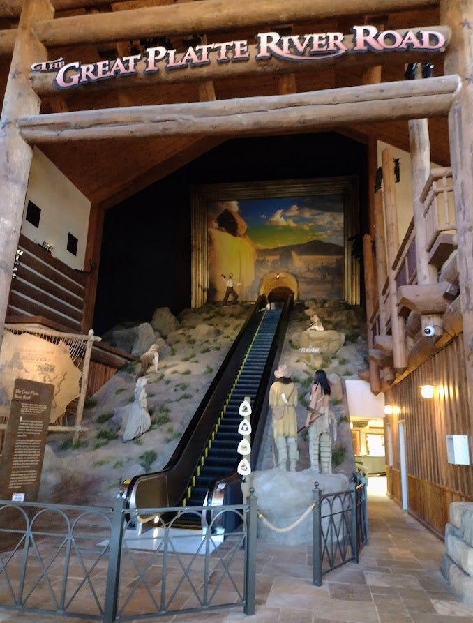 Escalator ending at mural of the road west, with mannequins of Indians and migrants watching or climbing