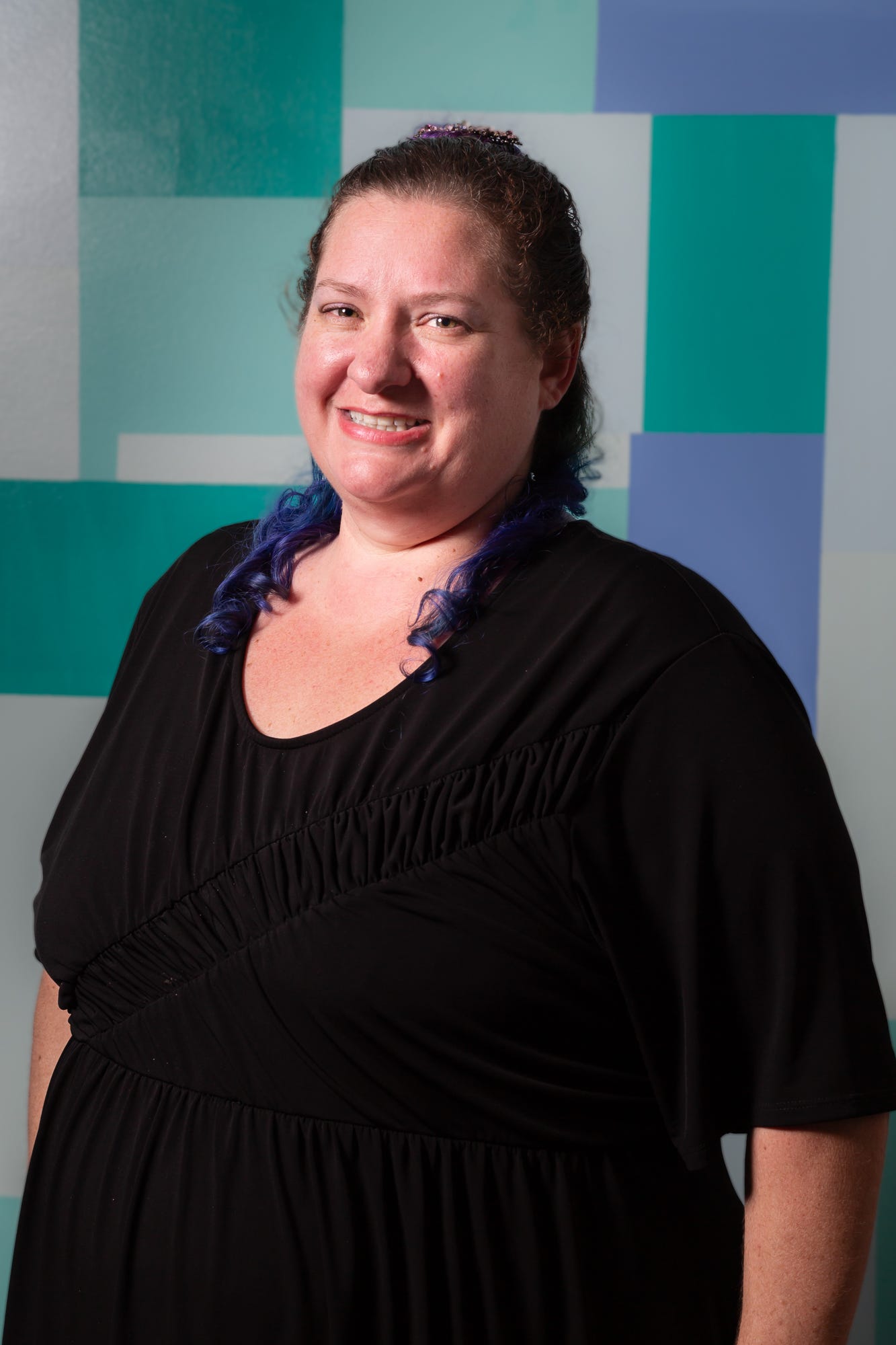 Portrait of a 40 year old, white skinned woman with light brown eyes smiles at the camera. She has dark brown, shoulder length, curly hair with blue tips, it is pulled back into a half ponytail. She is wearing a scoop neck black dress.