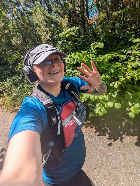 Selfie of Ellen running in a sunny woodland, she is wearing a cap, glasses and a Great North  Run 2024 t-shirt and waving at the camera and smiling.