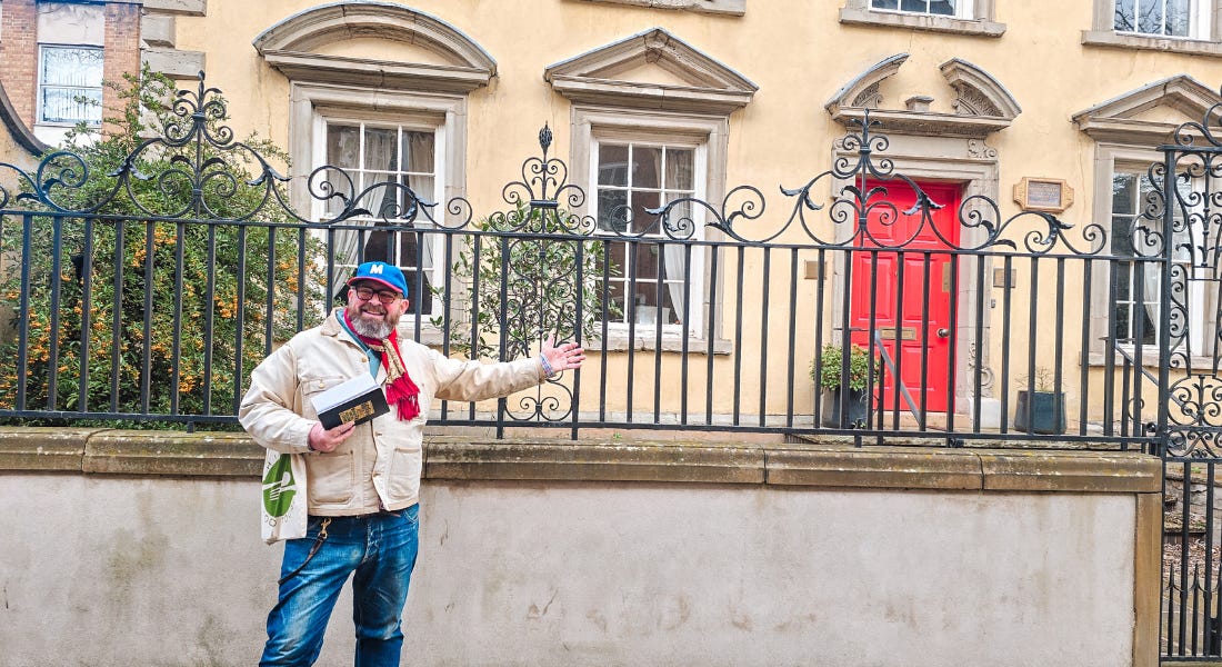 A man wearing a beige shirt and blue jeans standing in front of a large beige building with iron fencing