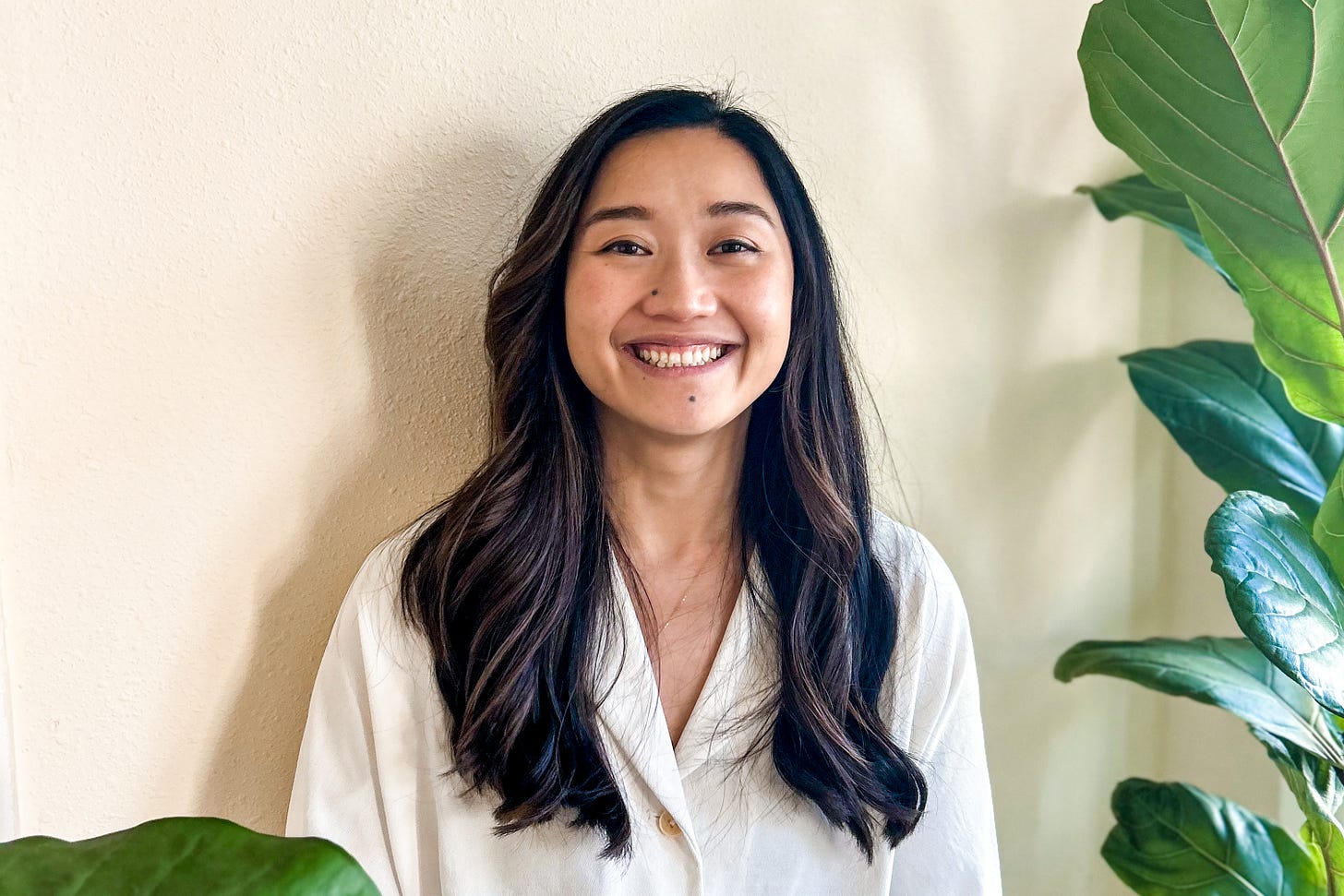 Asian American woman with wavy long brown hair surrounded by plants smiling at the camera