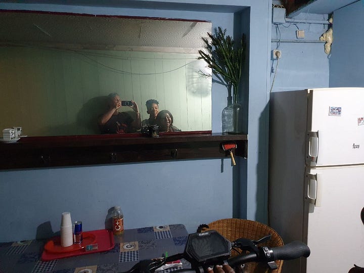 A mirror selfie of three people in front of a blue wall, there is a table and a fridge visible. And a poster with faces of different trans people, calling for a demonstration to honor the murdered Sara Millerey. 