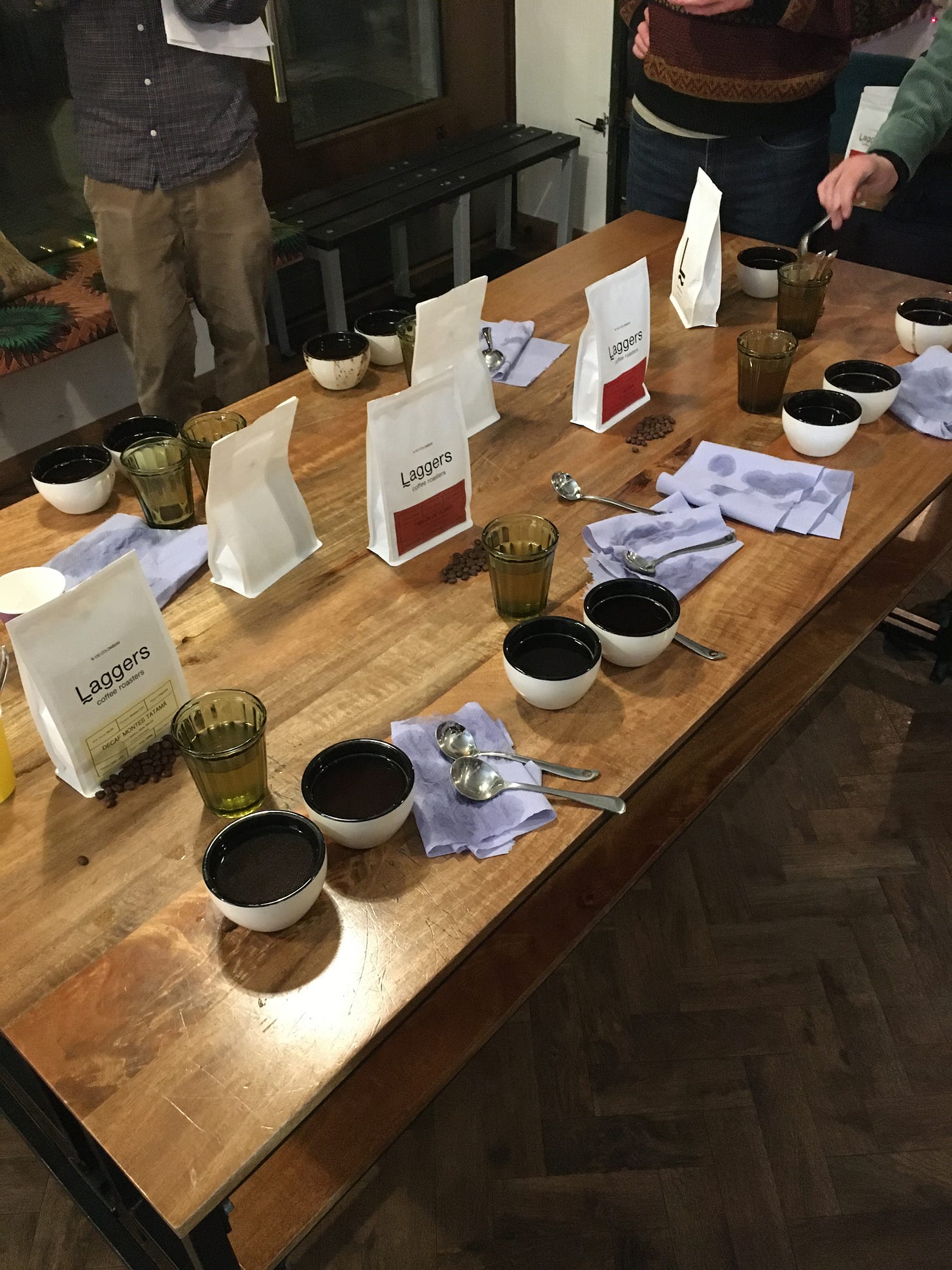 Wooden table with bags of coffee beans, glasses with water, cups of brewed coffee and spoons.