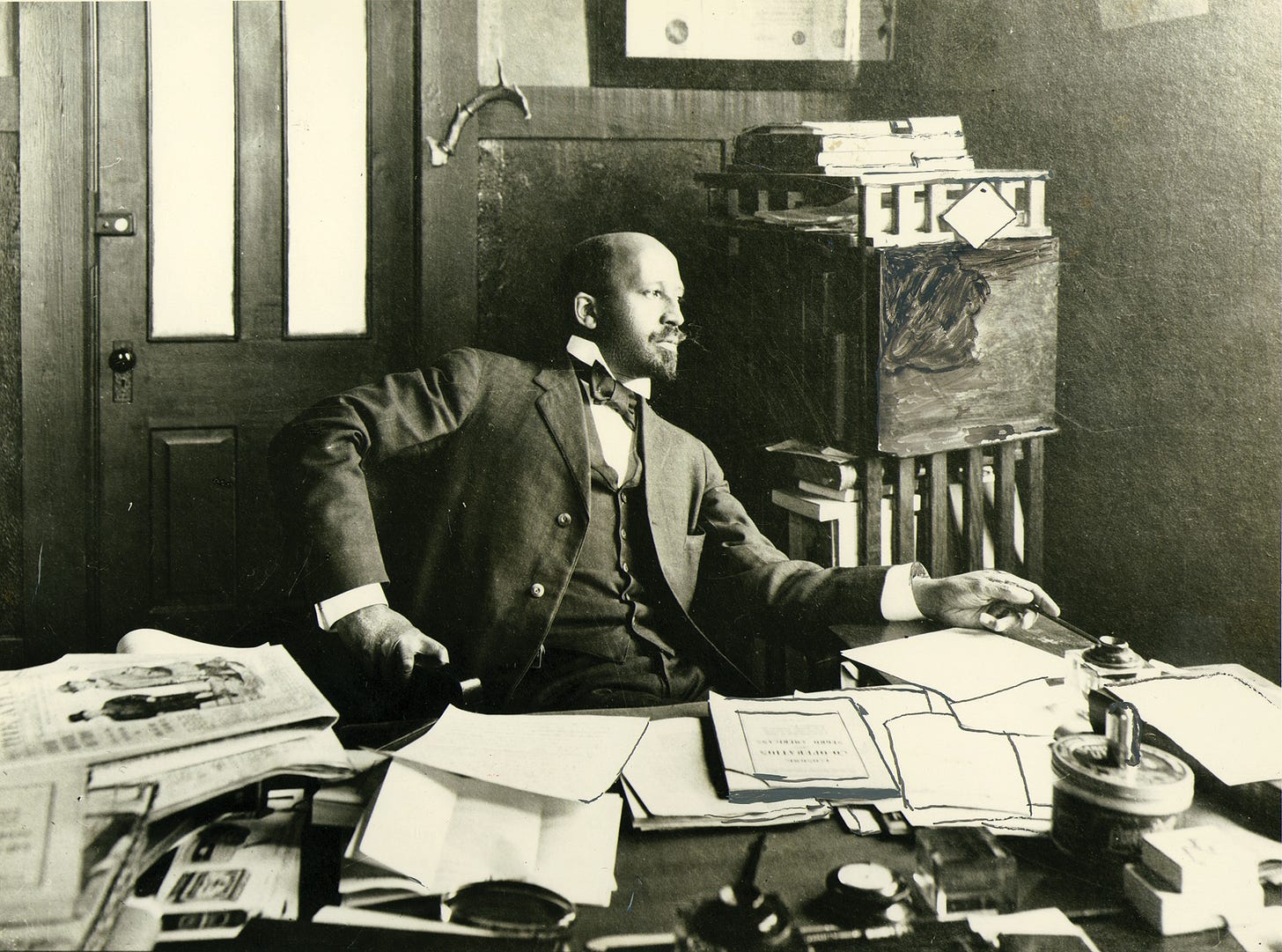  Black-and-white photograph of W.E.B. Du Bois seated at a cluttered desk in his office, wearing a three-piece suit with a bow tie. He leans back in his chair, looking thoughtfully to the side, with papers, books, and periodicals spread across the desk. The image captures Du Bois in a moment of intellectual focus and composure, surrounded by the tools of his scholarly and activist work.