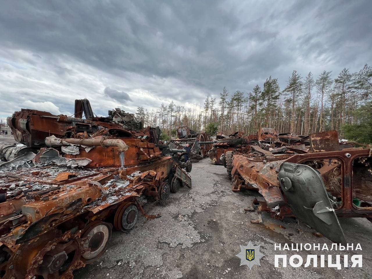 Rows of burned-out, rusted tanks and armored vehicles lined up on a road beside a pine forest under a cloudy sky. Rows of burned-out, rusted tanks and armored vehicles lined up on a road beside a pine forest under a cloudy sky.