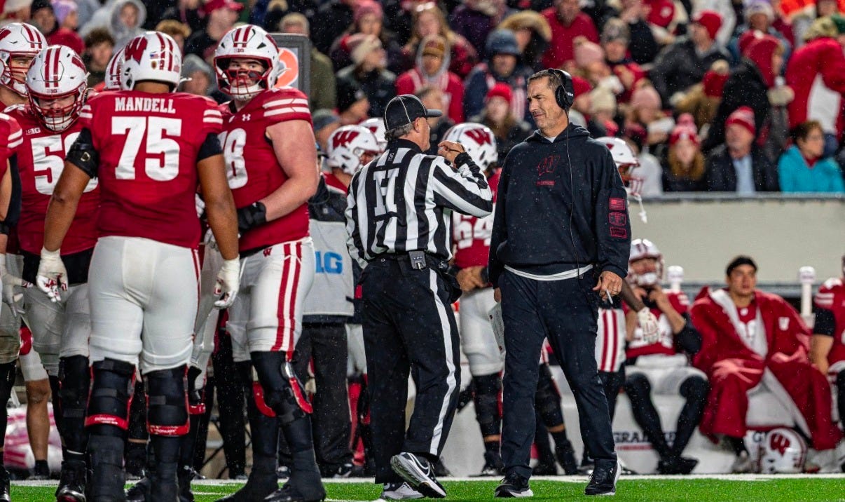 Wisconsin Badgers head coach Luke Fickell speaking with a game official as the offensive line stands nearby on the field.