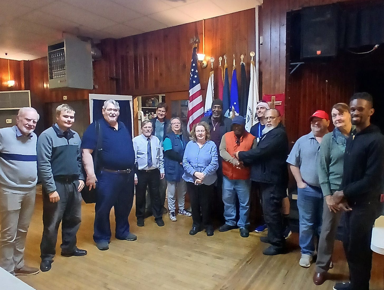 A group of about sixteen adults stands in a line for a group photo inside a wood‑paneled community hall. Behind them are the U.S. flag and several U.S. military service flags on stands, plus a red banner with a cross. The room has a hardwood floor, drop ceiling, speaker and small stage on the right, and wall-mounted HVAC units. The people wear casual to semi-formal clothing, including sweaters, button-down shirts, a tie, an orange vest, a red baseball cap, and a Santa hat, and they appear relaxed and smiling.