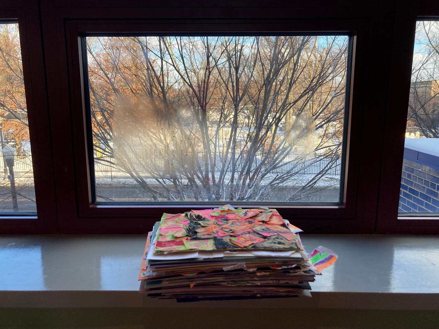 Dozens of collages stacked neatly on a shiny windowsill. The jagged bright scraps of paper that stick out from the pile are a nice contrast to the frosted window, bare branches, and snow outside.