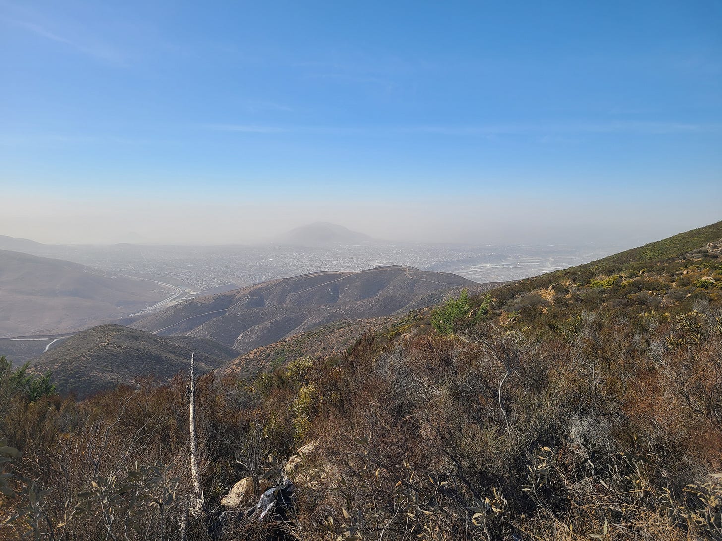a brush-filled mountain landscape with the border wall and Tijuana in the distance down the mountain a brush-filled mountain landscape with the border wall and Tijuana in the distance down the mountain