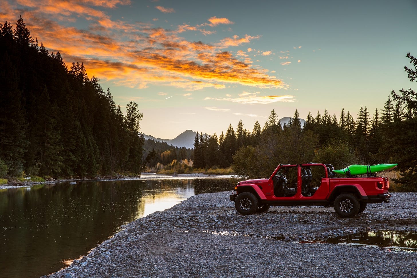 A red 2023 Jeep Gladiator Rubicon is parked next to a river at sunset with the roof and doors off and a kayak in the cargo bed.