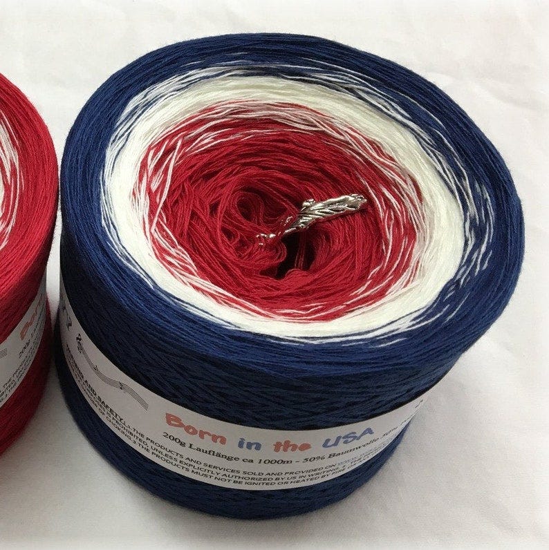 May include: A ball of red, white, and blue yarn with a label that reads 'Born in the USA 200g Lauflange ca 1000m - 30% Baumwolle'. The yarn is wound into a ball and is ready to be used for knitting or crocheting.