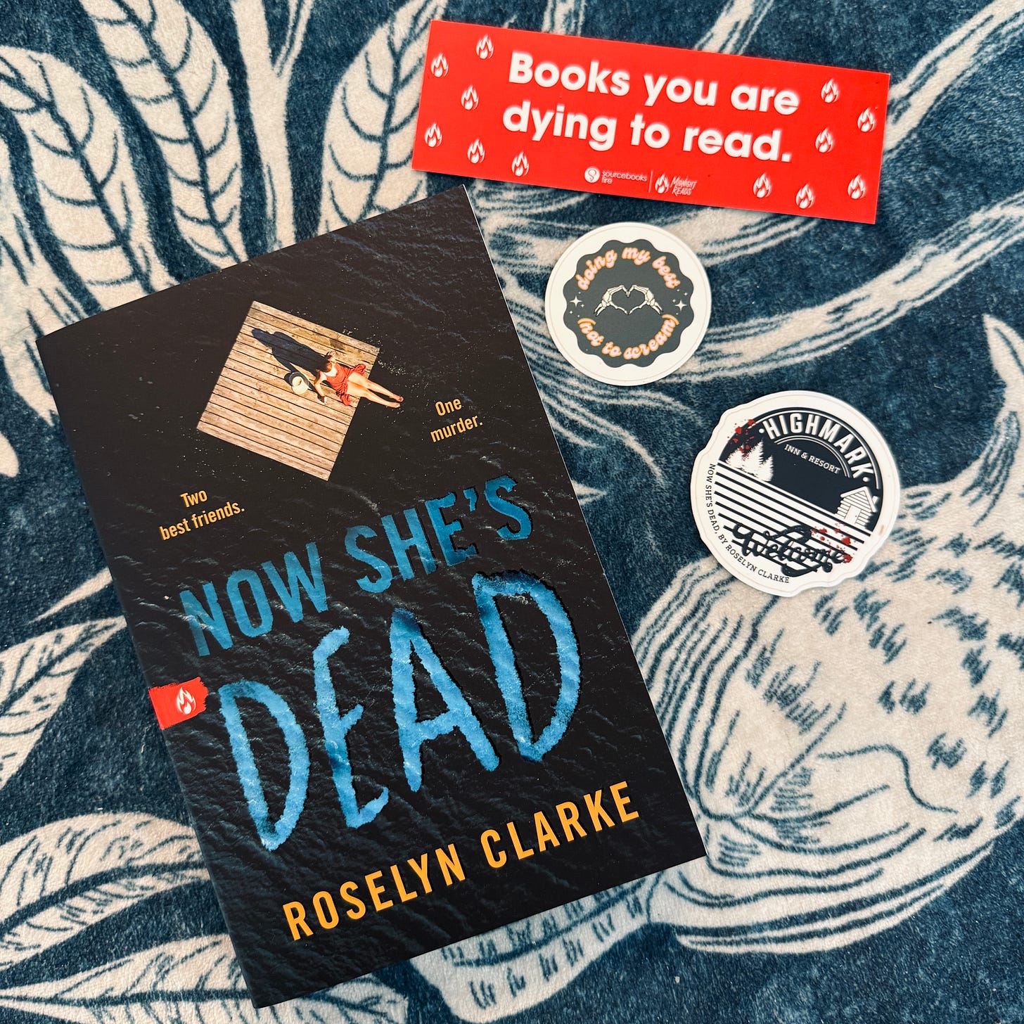 Copy of Now She's Dead next to a sticker with a Highmark Inn & Resort logo sticker and a sticker that says "Doing my Best (not to scream)." There's also a red bookmark with Sourcebooks Fire Midnight Reads branding that says "Books You Are Dying to Read."