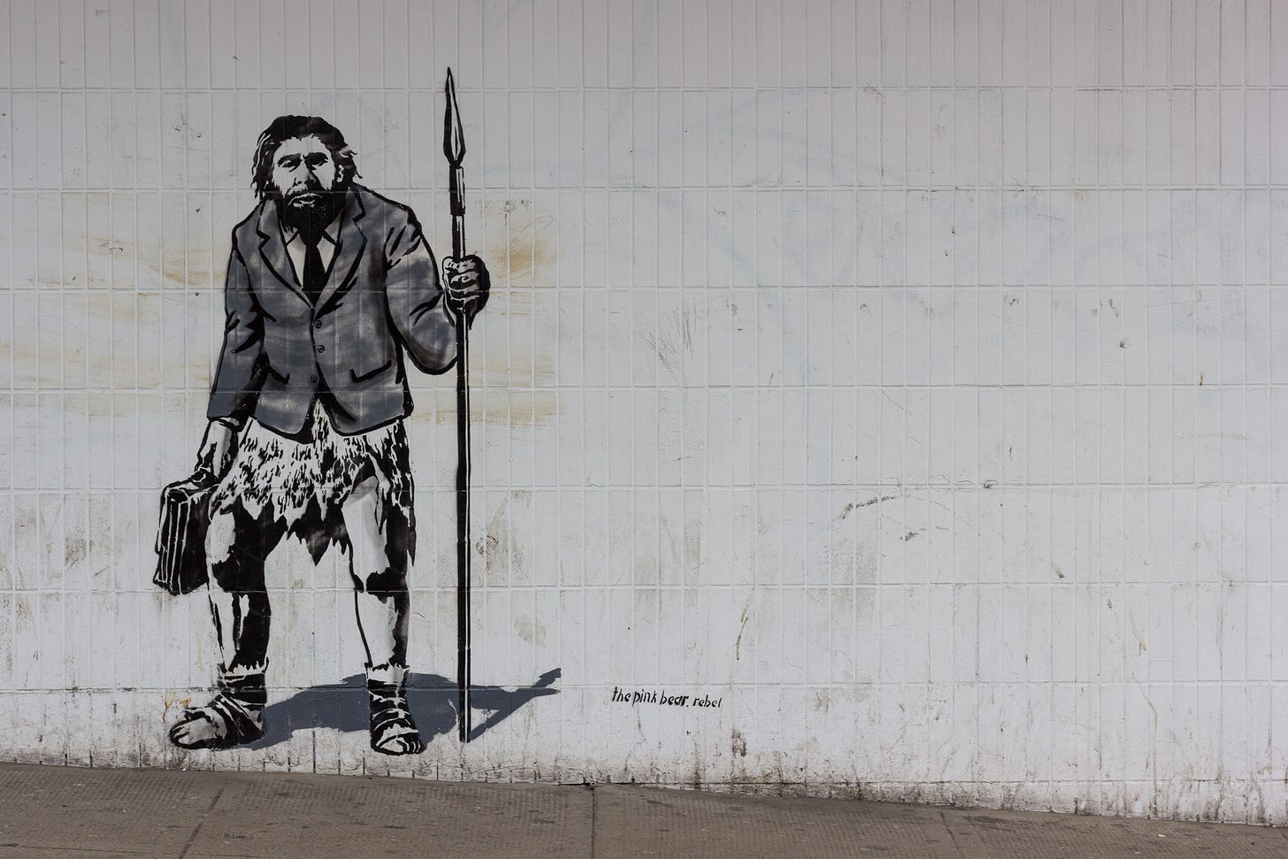 Graffiti in Schwarz und Grau auf einer weiß-grauen Gebäudewand. Es zeigt einen wohl männlichen Neandertaler. Am Oberkörper trägt er den Anzug eines Geschäftsmanns, ab der Hüfte eine Fellschürze. Die Beine sind nackt, an den Füßen und Schienbeinen trägt er Sandalenartiges. In der rechten Hand hält er einen Aktenkoffer, in der linken einen auf den Boden abgestützten Speer. Er schaut die Betrachter*innen an. Bild: Crawford Jolly / Unsplash Graffiti in Schwarz und Grau auf einer weiß-grauen Gebäudewand. Es zeigt einen wohl männlichen Neandertaler. Am Oberkörper trägt er den Anzug eines Geschäftsmanns, ab der Hüfte eine Fellschürze. Die Beine sind nackt, an den Füßen und Schienbeinen trägt er Sandalenartiges. In der rechten Hand hält er einen Aktenkoffer, in der linken einen auf den Boden abgestützten Speer. Er schaut die Betrachter*innen an. Bild: Crawford Jolly / Unsplash