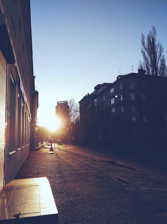 На зображенні вулиця, залита вранішнім світлом. Вдалині видно торговий центр, наполовину прихований яскравим сонцем. На передньому плані пуста вулиця та п'ятиповерхові будинки.