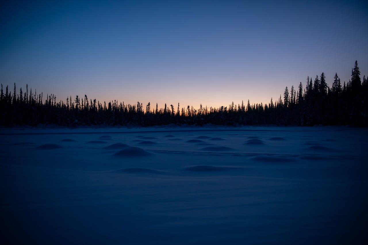 A wide frozen lake or pond stretches across the foreground under a deep blue twilight sky. A thin band of warm orange and peach light glows along the horizon behind the silhouetted treeline of black spruce. Ballaine Lake, Fairbanks, Alaska.