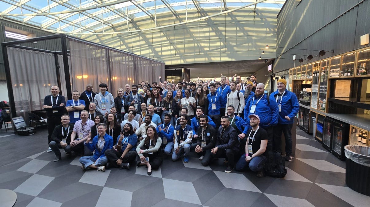 A large group gathered indoors for a conference or event, posing for a group photo. The setting includes a modern interior with a high glass ceiling, geometric floor tiles, a bar with bottle shelves, and a sectioned-off area with sheer curtains. Individuals wear lanyards, indicating a professional or social gathering.