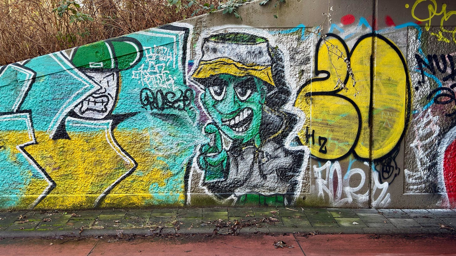 A vibrant graffiti mural features a green-faced character in a bucket hat, alongside various colorful abstract shapes and tags on a concrete wall. Duivendrecht, The Netherlands.