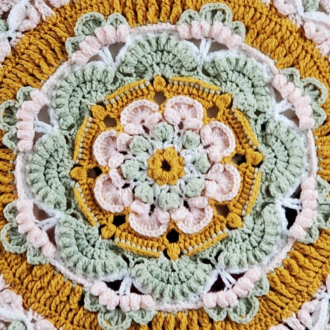 A crocheted mandala afghan blanket in goldenrod, sage, pink, and white.
