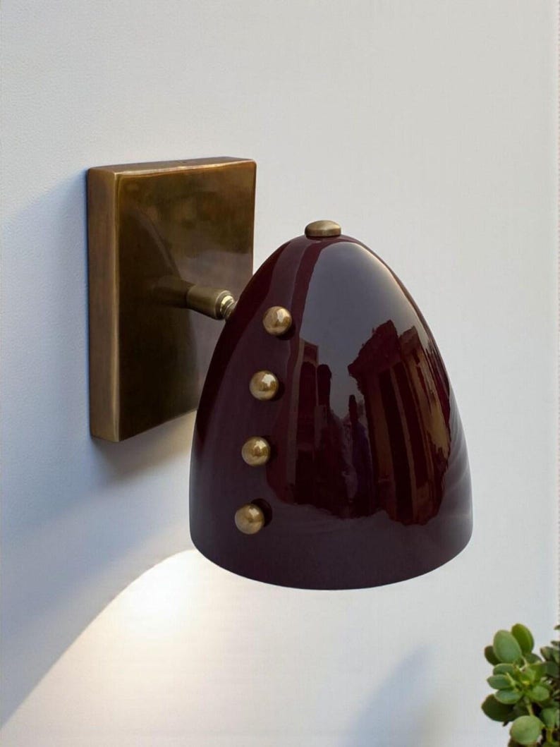 May include: A wall-mounted sconce with a burgundy shade and brass accents. The shade is a glossy, teardrop shape with three brass rivets. The sconce has a rectangular brass base and a small brass arm connecting the shade to the base. May include: A wall-mounted sconce with a burgundy shade and brass accents. The shade is a glossy, teardrop shape with three brass rivets. The sconce has a rectangular brass base and a small brass arm connecting the shade to the base.
