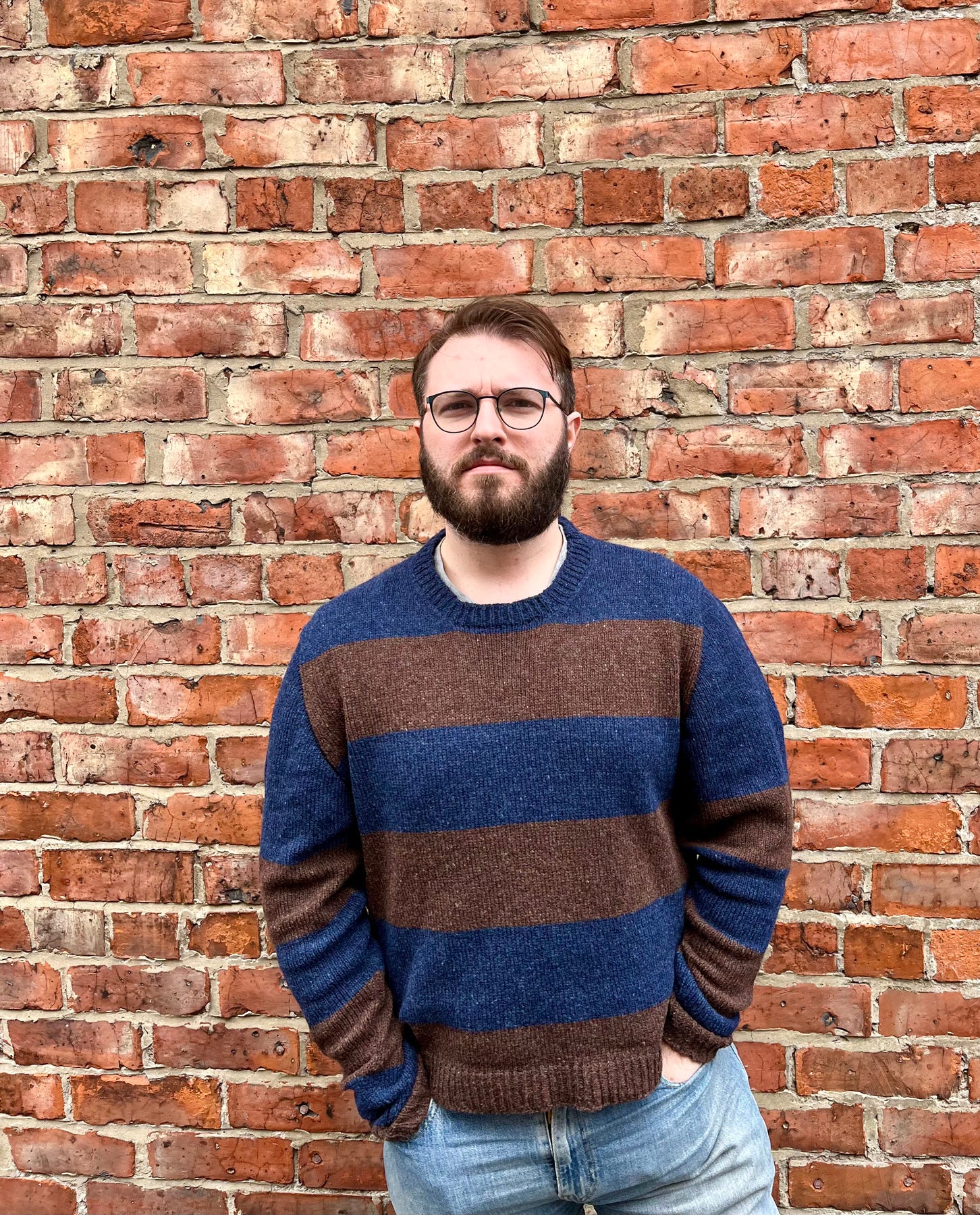 A man in his thirties wearing a wide-stripe chocolate and navy sweater standing in front of a red brick wall, hands in pockets, looking at the camera.