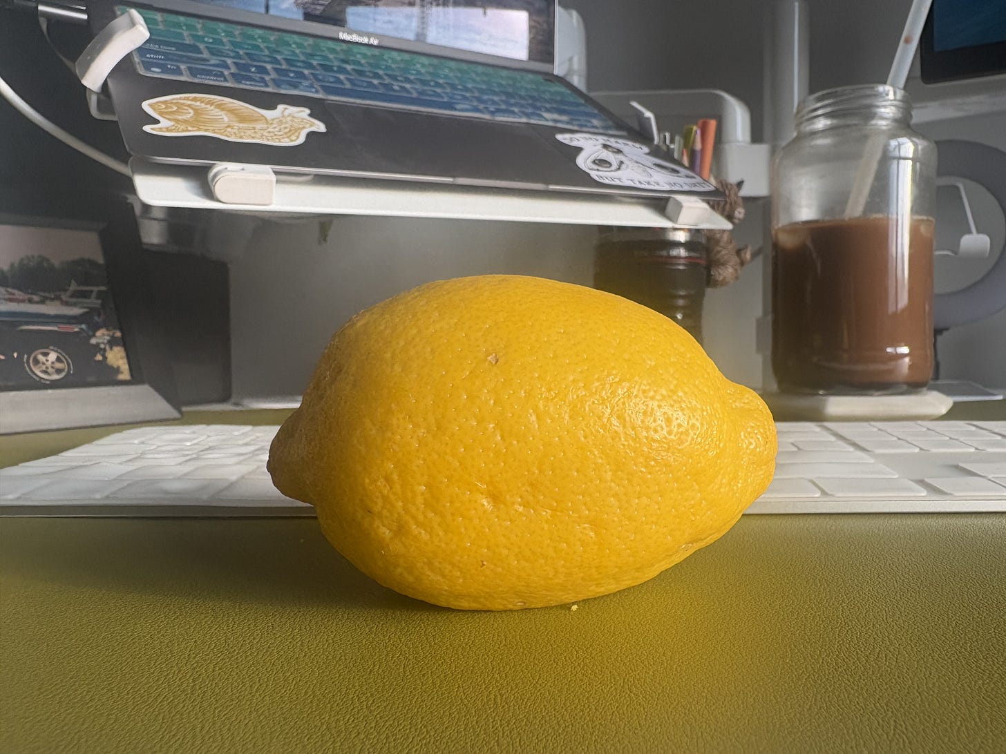 A whole bright yellow lemon rests on a green desk surface in front of a white keyboard and a raised laptop stand, with a glass jar of iced coffee blurred in the background. A whole bright yellow lemon rests on a green desk surface in front of a white keyboard and a raised laptop stand, with a glass jar of iced coffee blurred in the background.
