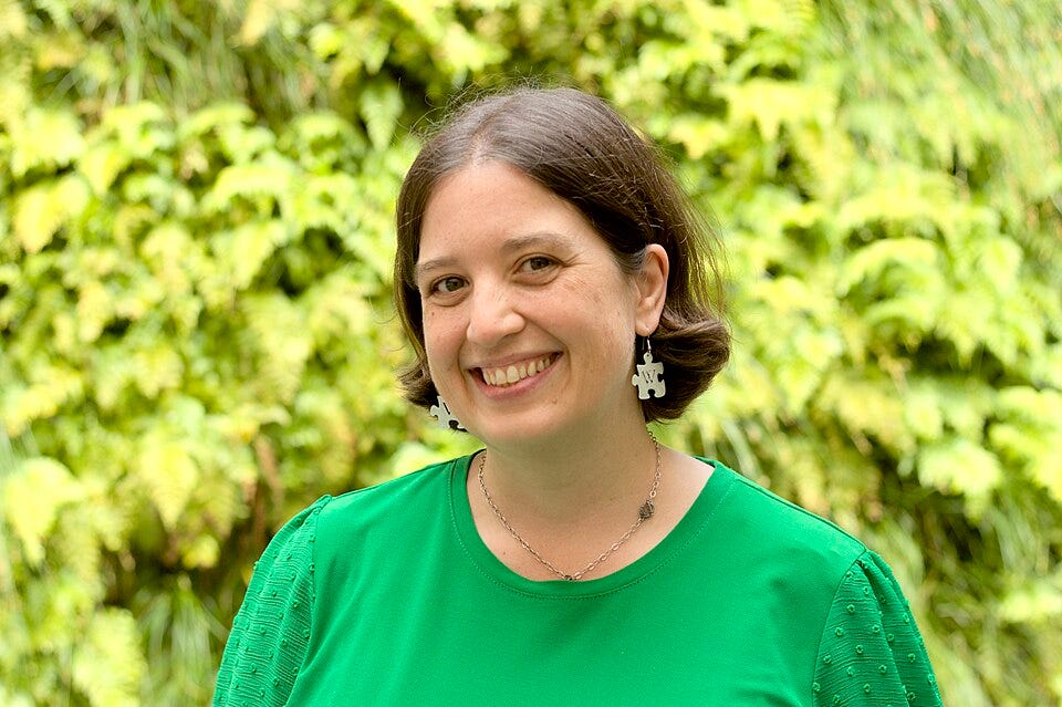 The portrait centers the woman from the chest up against a soft-focus background of green foliage. She wears a bright green blouse, a silver necklace, and white puzzle-piece earrings marked with a “W,” with her face and upper body in sharp focus.