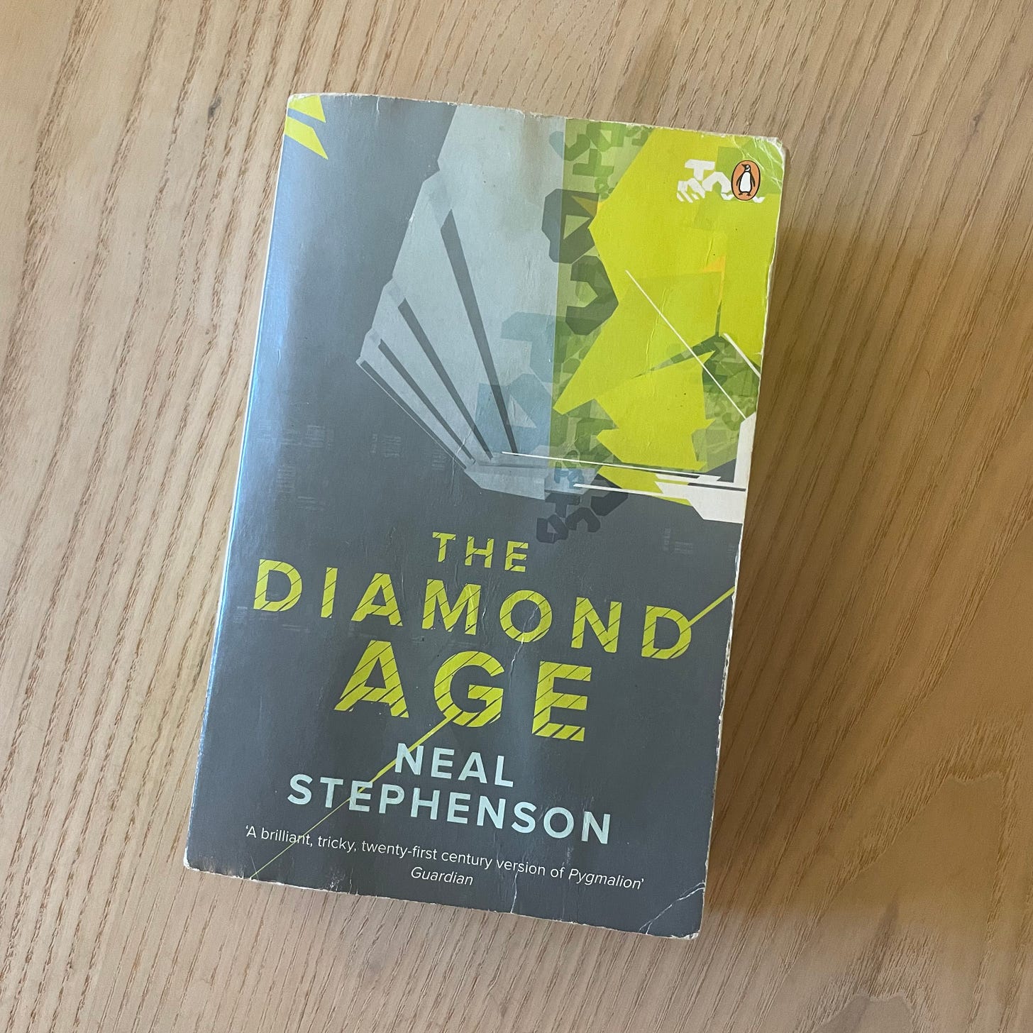 Paperback copy of 'The Diamond Age' by Neal Stephenson, published by Penguin. The cover features a minimalist design with the title in bright yellow-green text against a dark gray background, with abstract geometric shapes suggesting mechanical or technological elements. A Guardian review quote calls it 'A brilliant, tricky, twenty-first century version of Pygmalion'