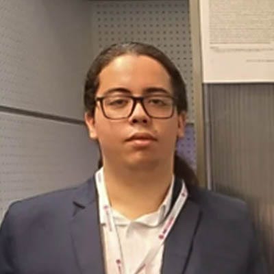 foto de um homem com cabelos compridos presos em um rabo de cavalo. Ele usa óculos, veste uma camisa branca e um blazer cinza chumbo.