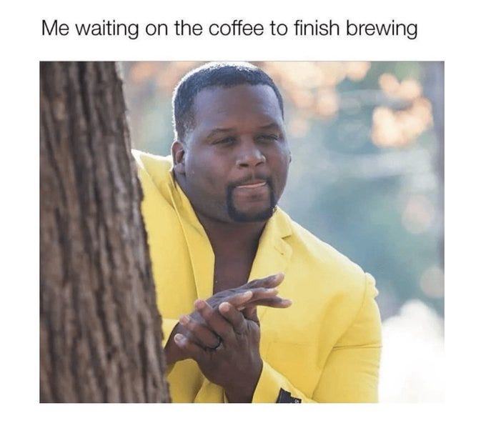 A Black man with a beard wearing a yellow suit jacket and shirt stands behind a tree trunk in an outdoor setting with blurred green foliage in the background. He leans forward with hands clasped in front of him, looking directly at the viewer with a patient yet anticipatory facial expression. The image is formatted as a meme with overlaid white text at the top reading Me waiting on the coffee to finish brewing.