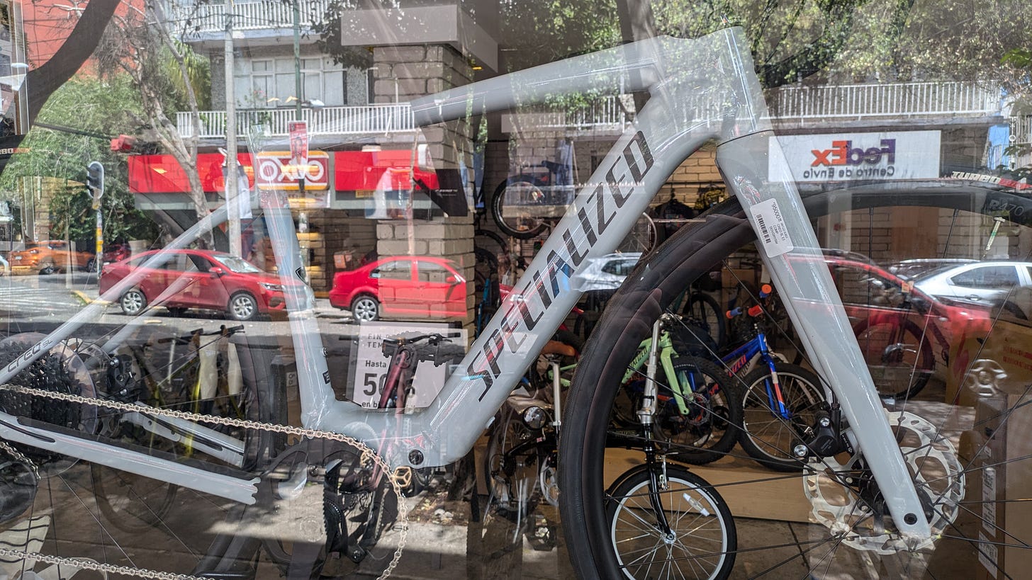 A Specialized bicycle displayed behind a bike shop window, with reflections of passing cars, trees, and an OXXO across the street.