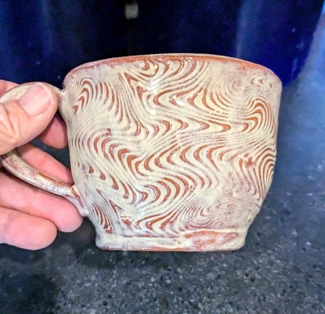 May include: A handmade ceramic mug with a unique swirl pattern in cream and reddish-brown. The mug has a rounded shape with a handle, perfect for coffee or tea. The mug is held by a person.