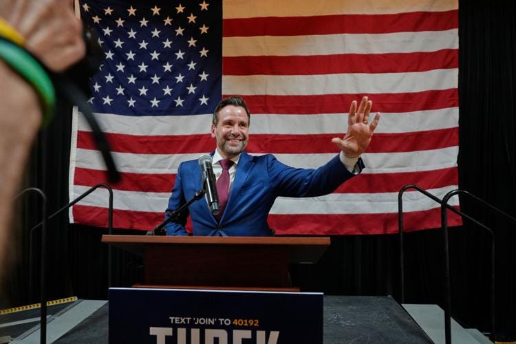 A white man is smiling and waving from a podium concealing his manual wheelchair in front of a large American flag. A white man is smiling and waving from a podium concealing his manual wheelchair in front of a large American flag.