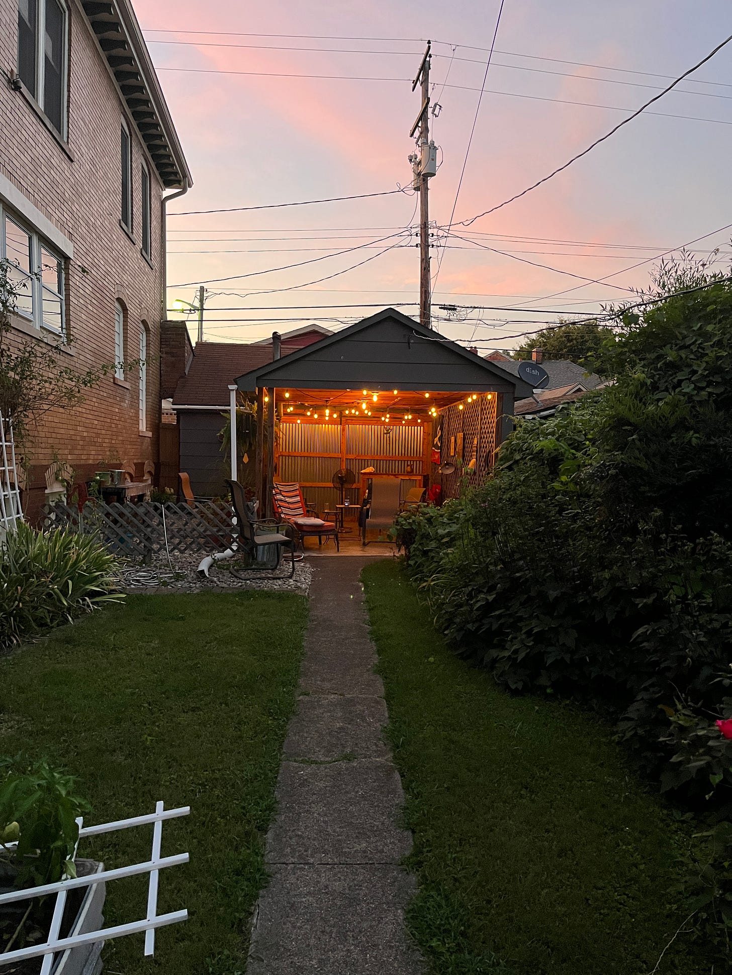 View into our backyard, with lots of vines and tomato plants and then the party pavilion lit up with fairy lights and above the sky is pink and light blue