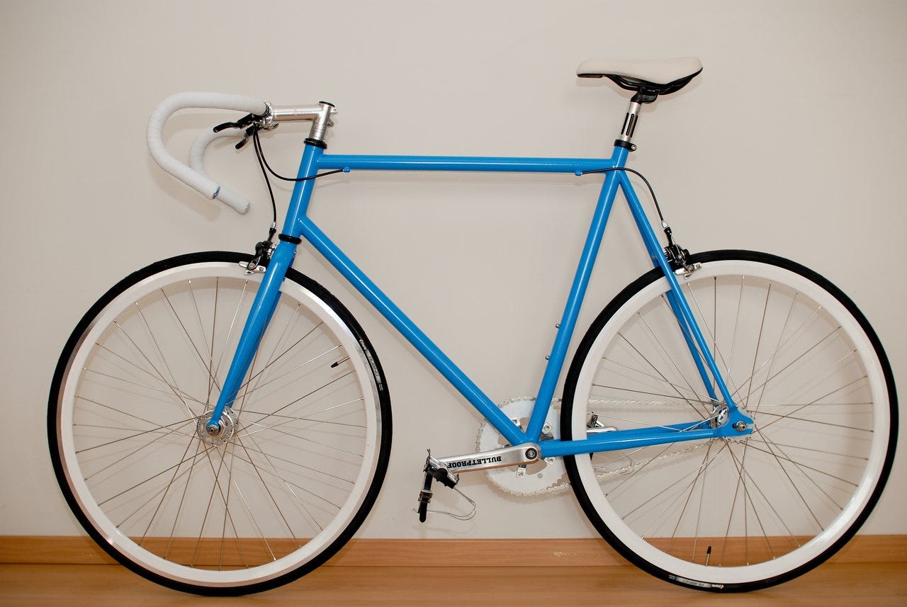 A blue road bike with white handlebars and wheels leans against a plain wall indoors. A blue road bike with white handlebars and wheels leans against a plain wall indoors.