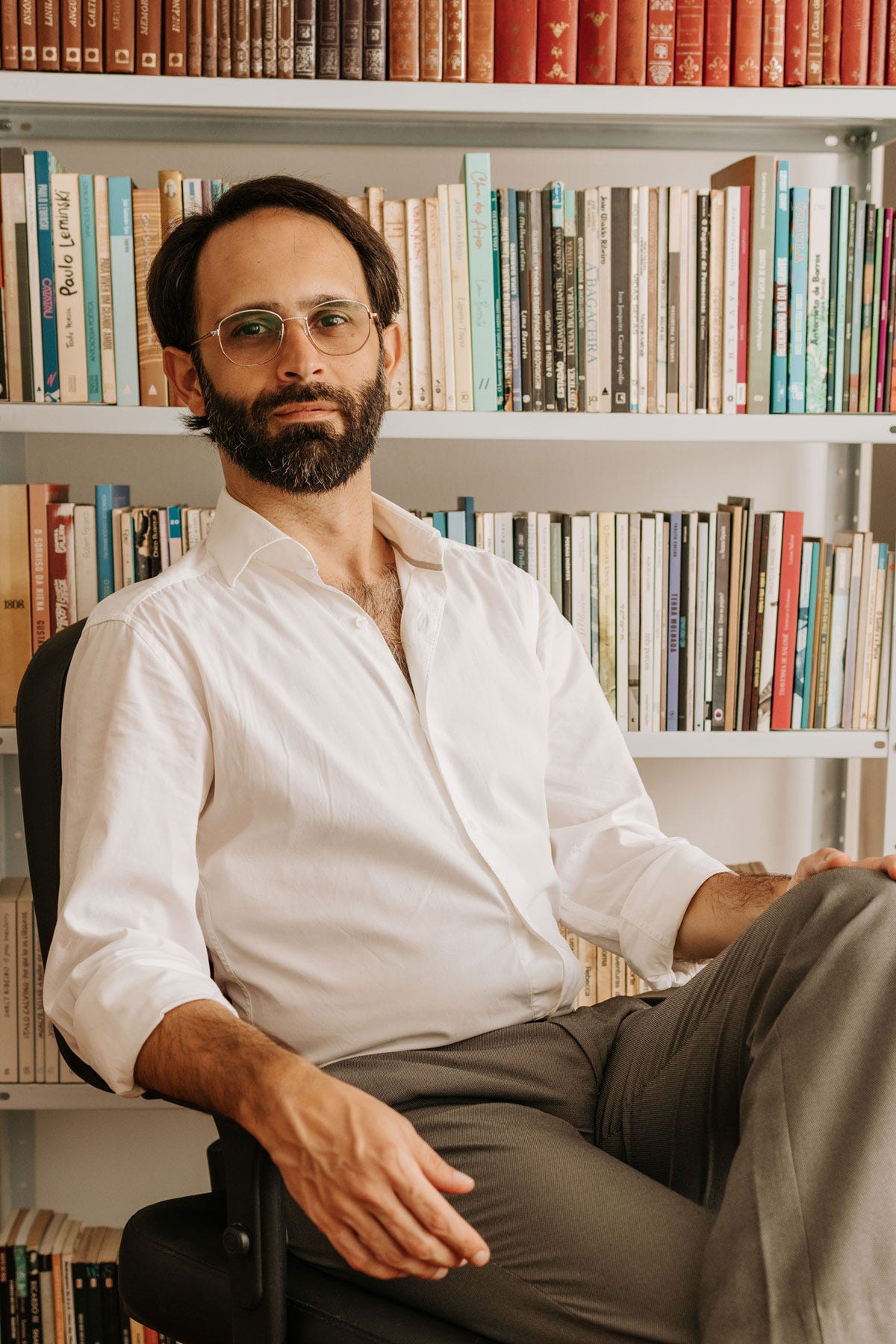 Foto de Christian von Koenig, um escritor de 35 anos à frente de uma estante cheia de livros em casa. Estou sentado confortável numa cadeira de escritório. Tenho a pele branca bronzeada, cabelo e barba castanhos com pelos brancos, uso óculos redondos e visto uma camisa social branca com uma calça social cinza.