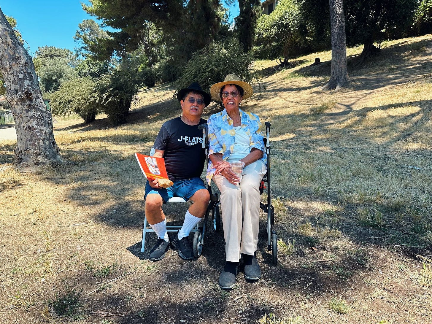 an elder japanese american man wearing a shit that says "J-Flats" sitting next to an elder Black woman in a light blue shirt. in the background is a park with dry grass and trees