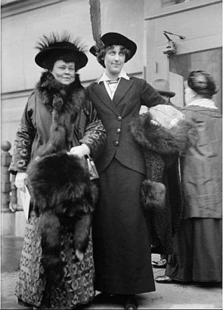 A fancy dressed women with a fur hat and coat on, arm in arm with another women, dressed less fancily, but still in a frock and dress and hat. A fancy dressed women with a fur hat and coat on, arm in arm with another women, dressed less fancily, but still in a frock and dress and hat.
