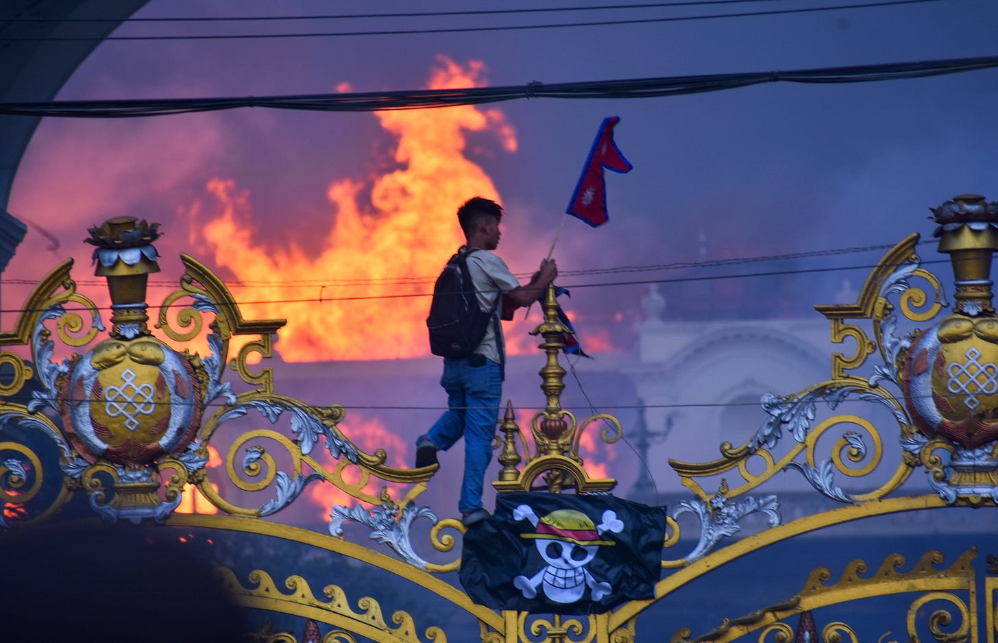 A protester carrying a Nepalese flag hangs a pirate flag on an ornate gate, as smoke and flames rise from a palace building in the background. A protester carrying a Nepalese flag hangs a pirate flag on an ornate gate, as smoke and flames rise from a palace building in the background.
