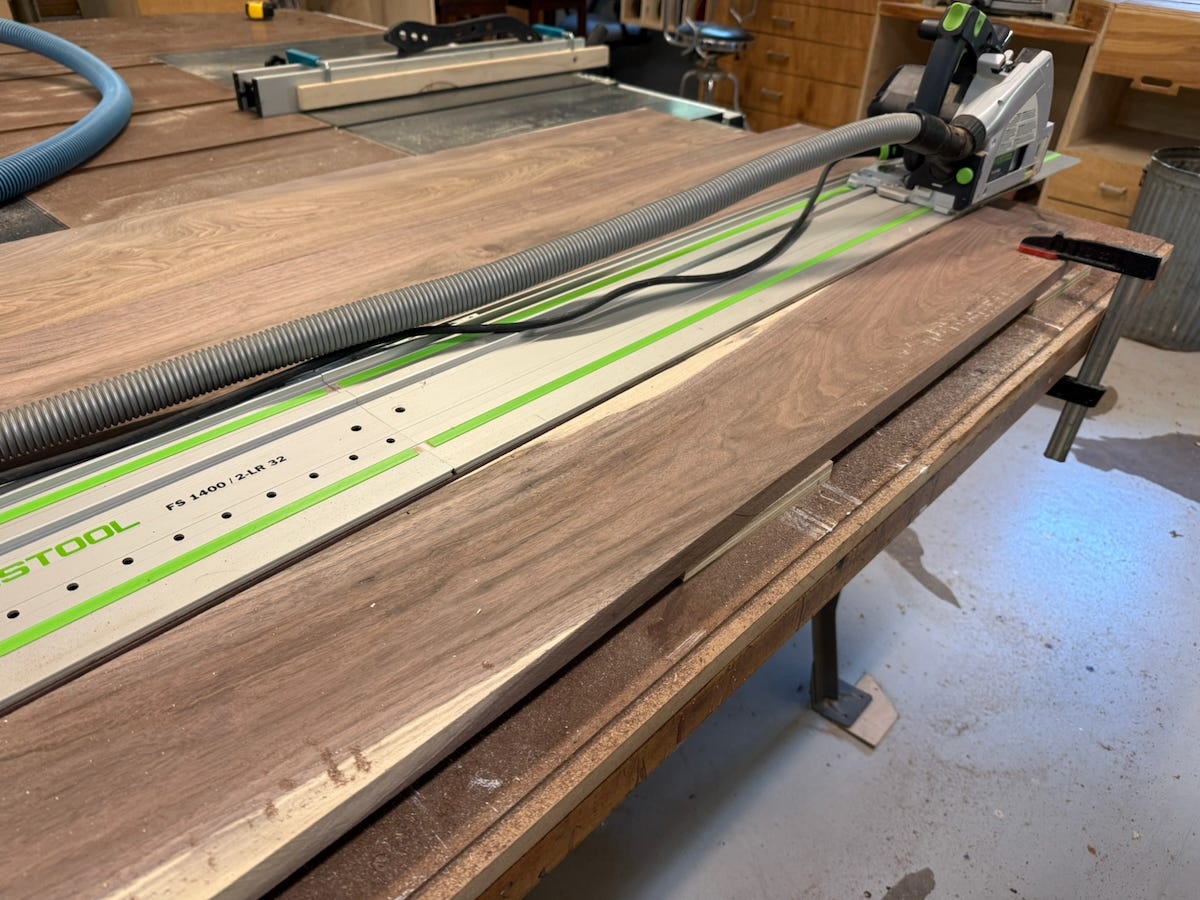 A woodworking shop scene showing a Festool track saw cutting a long walnut board on a workbench. The saw rides along an aluminum guide rail connected to a dust-extraction hose, allowing for a precise straight cut. Clamps secure the workpiece to the bench, and fine sawdust is visible around the cut line. Additional woodworking tools and benches can be seen in the background.