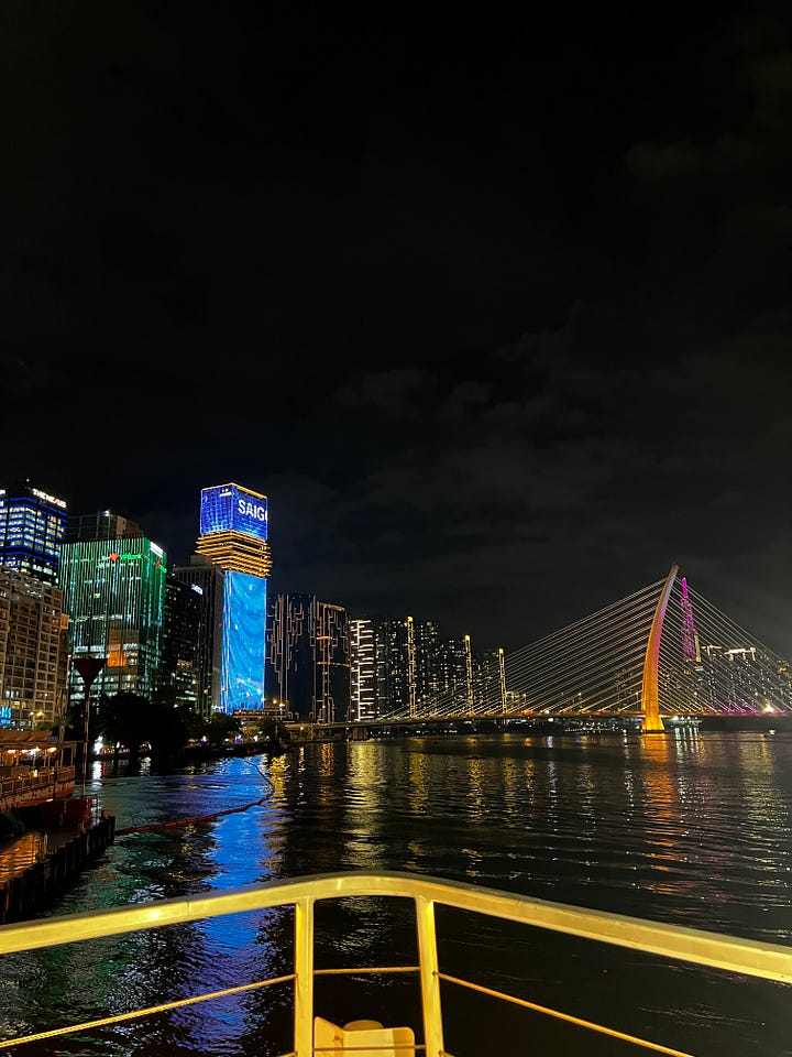 Nighttime view of Ho Chi Minh City skyline along the Saigon River, with illuminated high-rises—including a tower lit in red with yellow stars and another displaying “SAIGON”—and a glowing cable-stayed bridge, reflected in the water from a boat railing in the foreground.