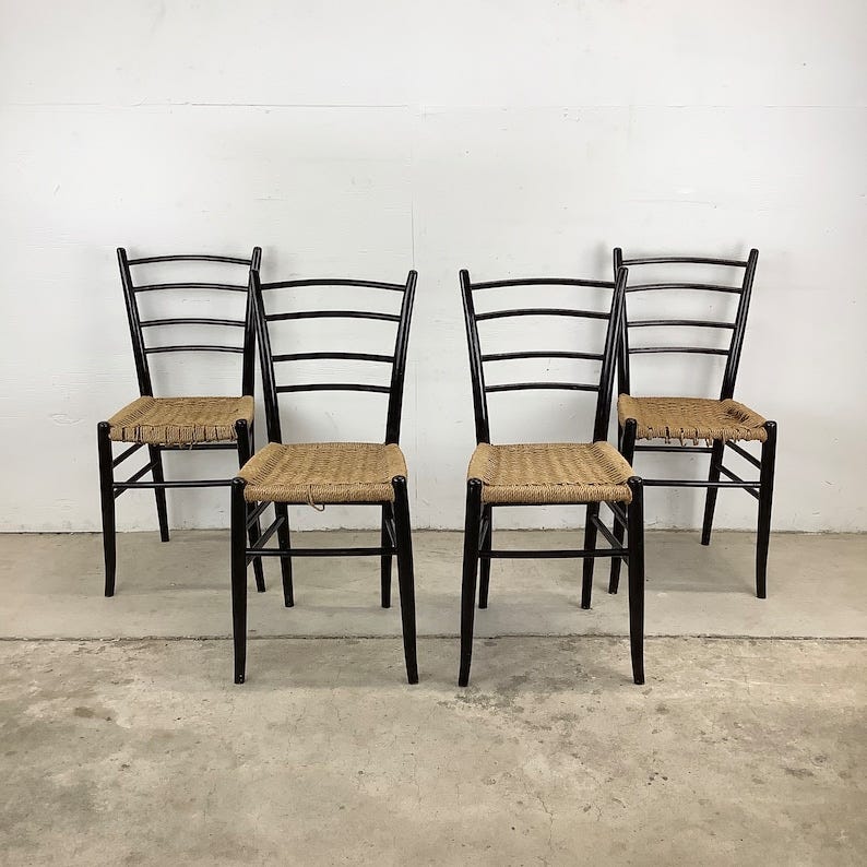 May include: Four black chairs with woven cane seats. The chairs are arranged in a row, with the backs of the chairs facing the viewer. The chairs are all identical in design and have a simple, elegant style.