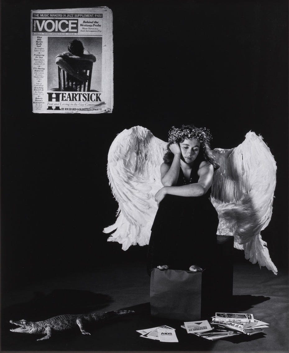 A woman wearing large, white angel wings, and a flower crown, sits barefoot pensively on a box. There's a newspaper with David Wojnarowicz on the wall behind her and a small faux alligator on the ground in front of her along with several AIDS and safe sex pamphlets, creating a surreal scene. The photo is in black and white.