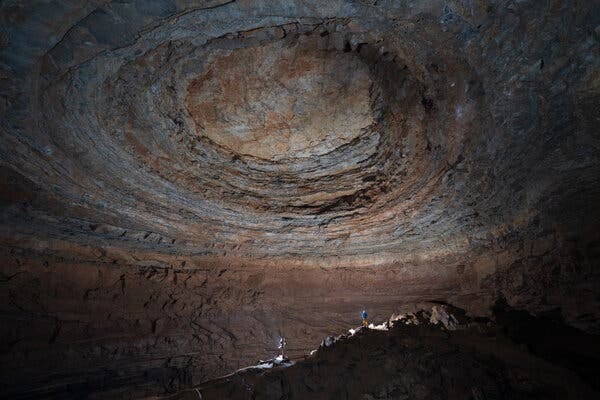 Deep in a cave, two people stand inside a vast underground space with a circular pattern in the cavern ceiling, far above them.