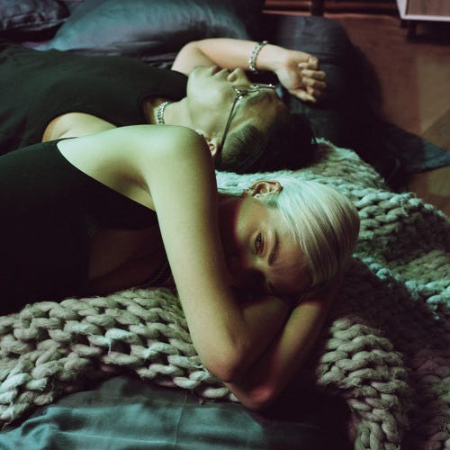 “Two people lie side by side on a bed covered with a chunky knit blanket, bathed in moody green-toned lighting. In the foreground, a person with short platinum-blonde hair rests their head on their arm, looking toward the camera with a calm, distant expression. Behind them, another person with dark hair pulled back and wearing glasses reclines with an arm draped over their head. Both wear black clothing and subtle jewelry. The atmosphere feels intimate and introspective, with soft textures and low light. A red ‘Parental Advisory’ label sits in the lower right corner. “Two people lie side by side on a bed covered with a chunky knit blanket, bathed in moody green-toned lighting. In the foreground, a person with short platinum-blonde hair rests their head on their arm, looking toward the camera with a calm, distant expression. Behind them, another person with dark hair pulled back and wearing glasses reclines with an arm draped over their head. Both wear black clothing and subtle jewelry. The atmosphere feels intimate and introspective, with soft textures and low light. A red ‘Parental Advisory’ label sits in the lower right corner.